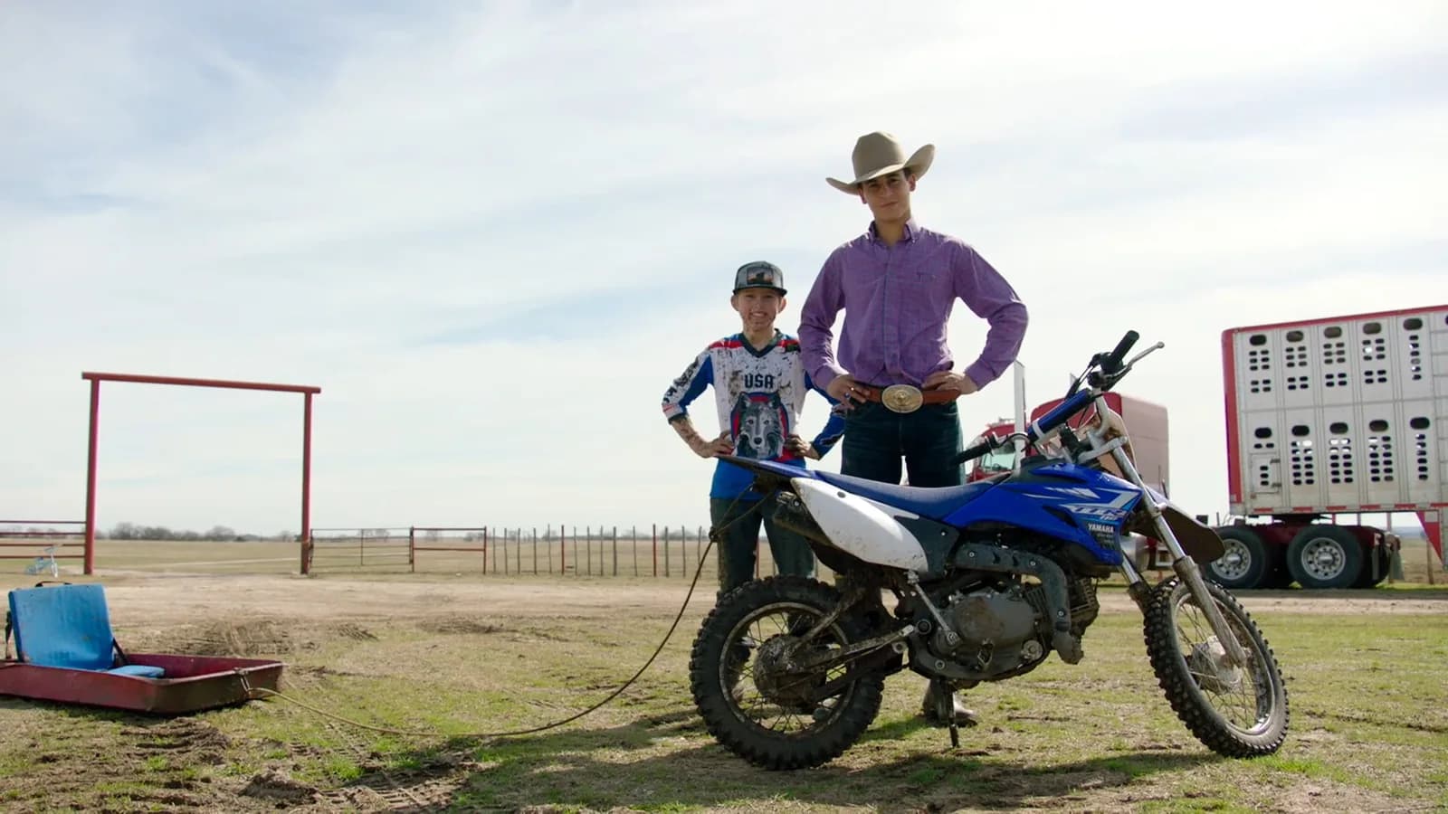 Two young people standing behind a blue dirt bike on a rural dirt lot with a livestock trailer in the background