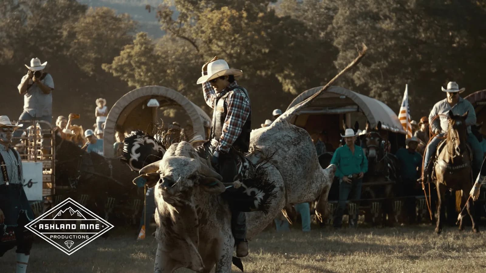 Cowboy riding a bucking bull at a rodeo with spectators and covered wagons, Ashland Mine logo watermark