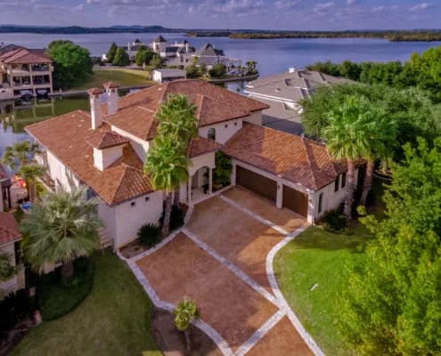 Aerial view of a two-story house with red tile roof, curved brick driveway, palm trees, and nearby water
