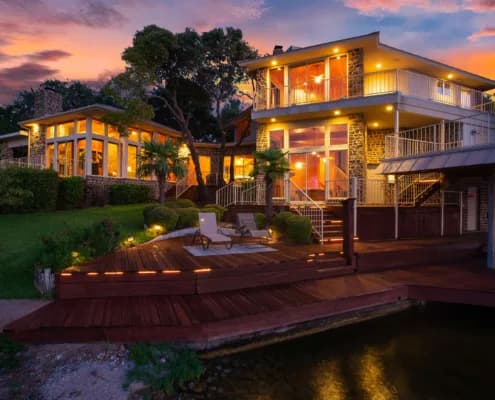 Two-story house with large glass windows and warm lights, wooden deck by the water at sunset