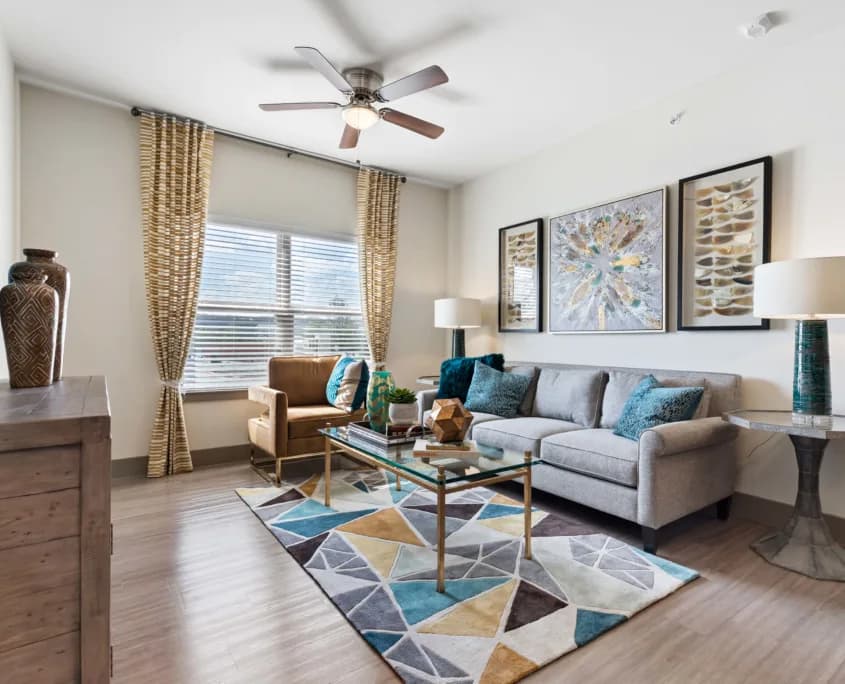 Living room with gray sofa and teal cushions, glass coffee table, geometric rug, armchair, wall art, lamps
