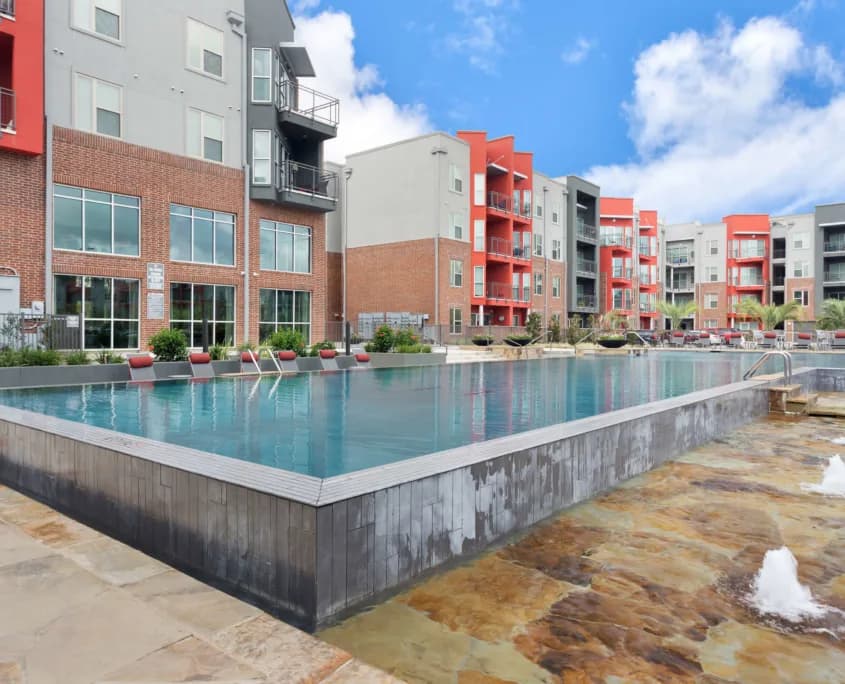 Rectangular pool with modern brick and gray apartment buildings, red balconies, under a blue sky