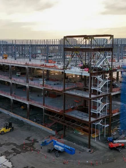 Steel-framed multi-story building under construction with exposed floors, stairs, and construction vehicles