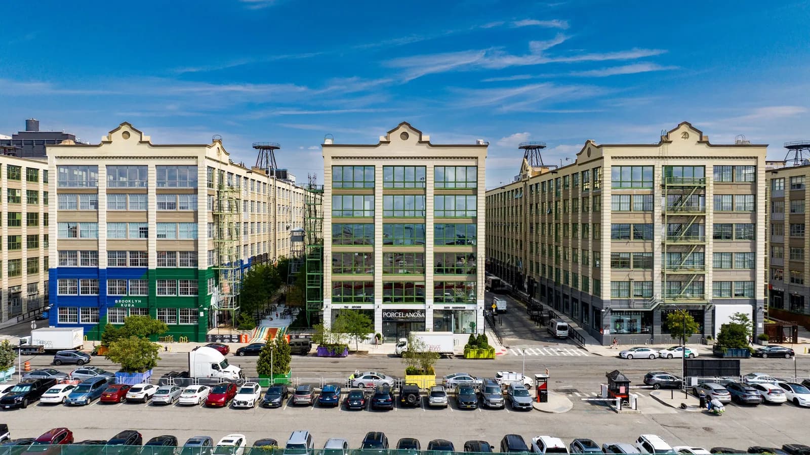 Three beige midrise buildings with large glass windows, foreground row of parked cars, blue sky