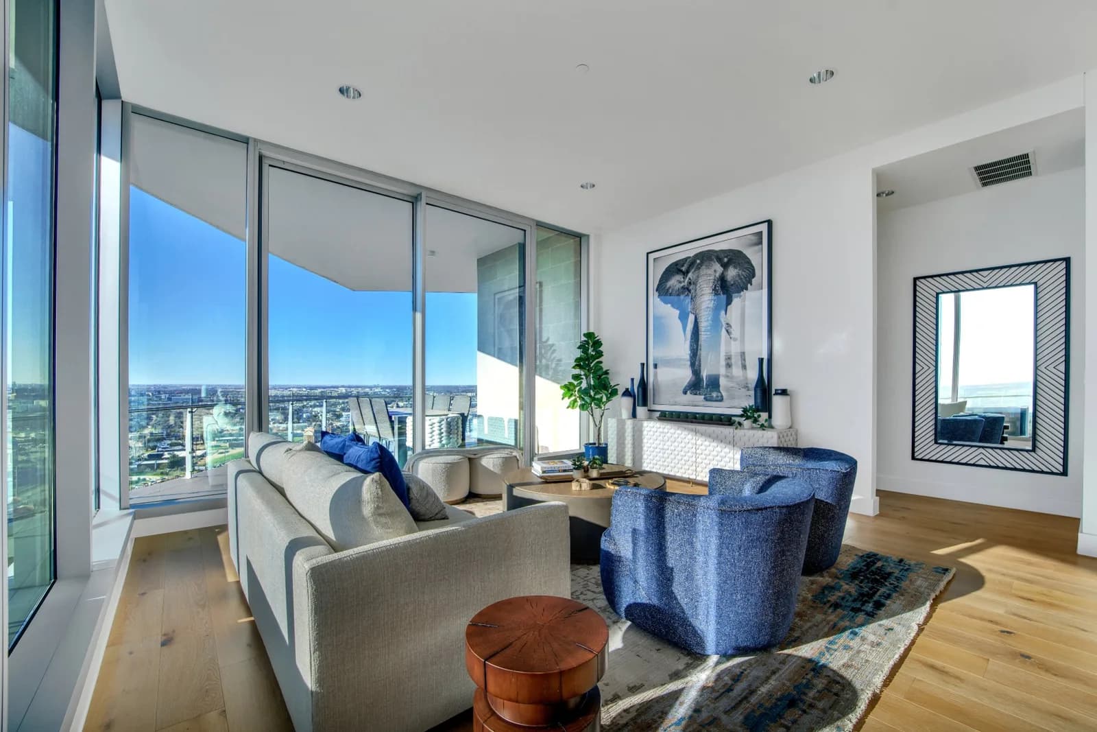 Living room with floor-to-ceiling windows and city view, beige sofa, blue chairs, plant, elephant photograph