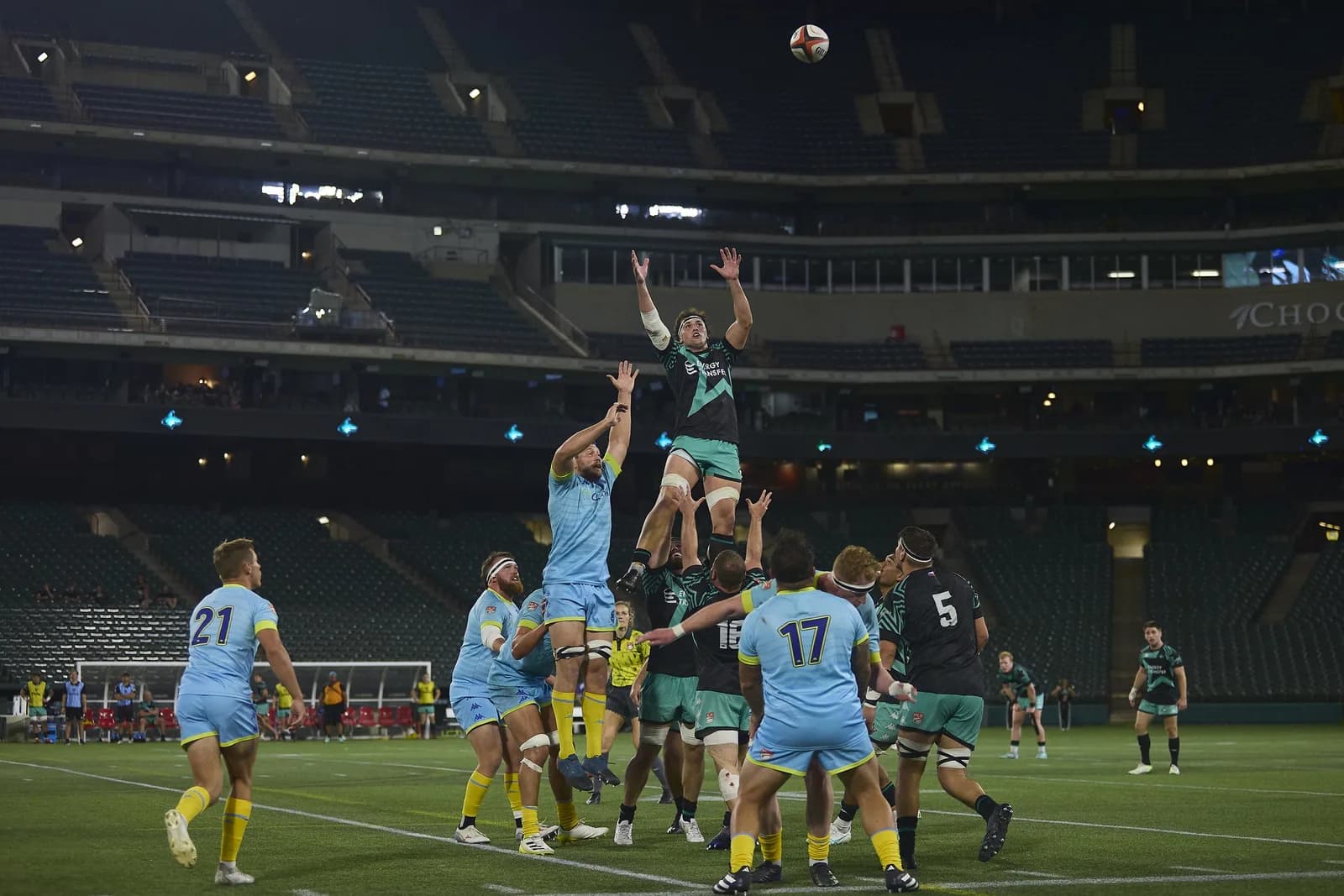 Rugby players in teal and light-blue jump for a lineout as the ball rises above them in the stadium