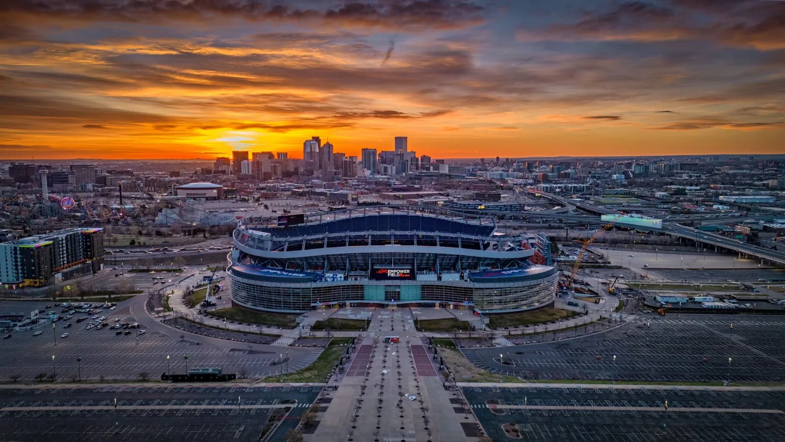 Aerial view of a large curved stadium at sunset with a city skyline in the distance