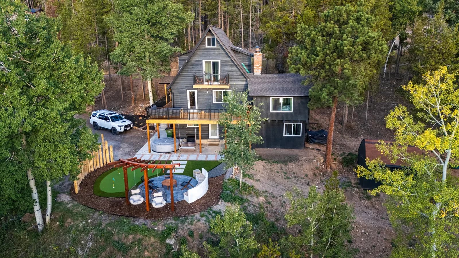 Two-story dark-gray house with balcony; white SUV on gravel driveway; pergola seating and mini golf