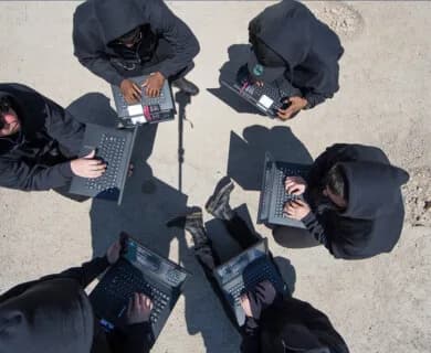 Top-down view of seven people in dark hoodies using laptops on a sandy surface