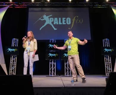 Stage with two presenters, woman with mic and man with open arms, paleo fx logo on screen