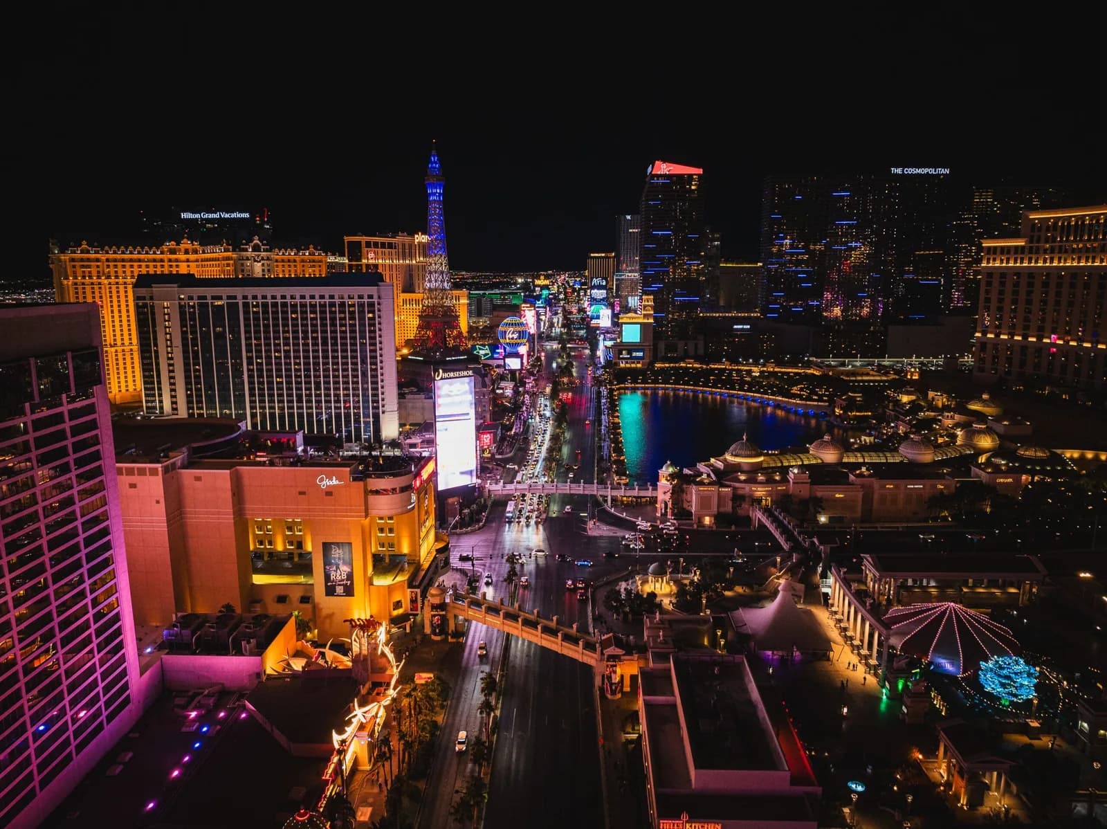 Night aerial view of a brightly lit city along a river, with tall hotels, neon signs, and a blue-lit tower