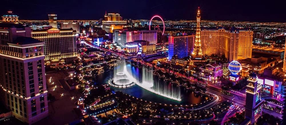 Night aerial view of a neon-lit city with a circular fountain, a pink Ferris wheel, and tall buildings