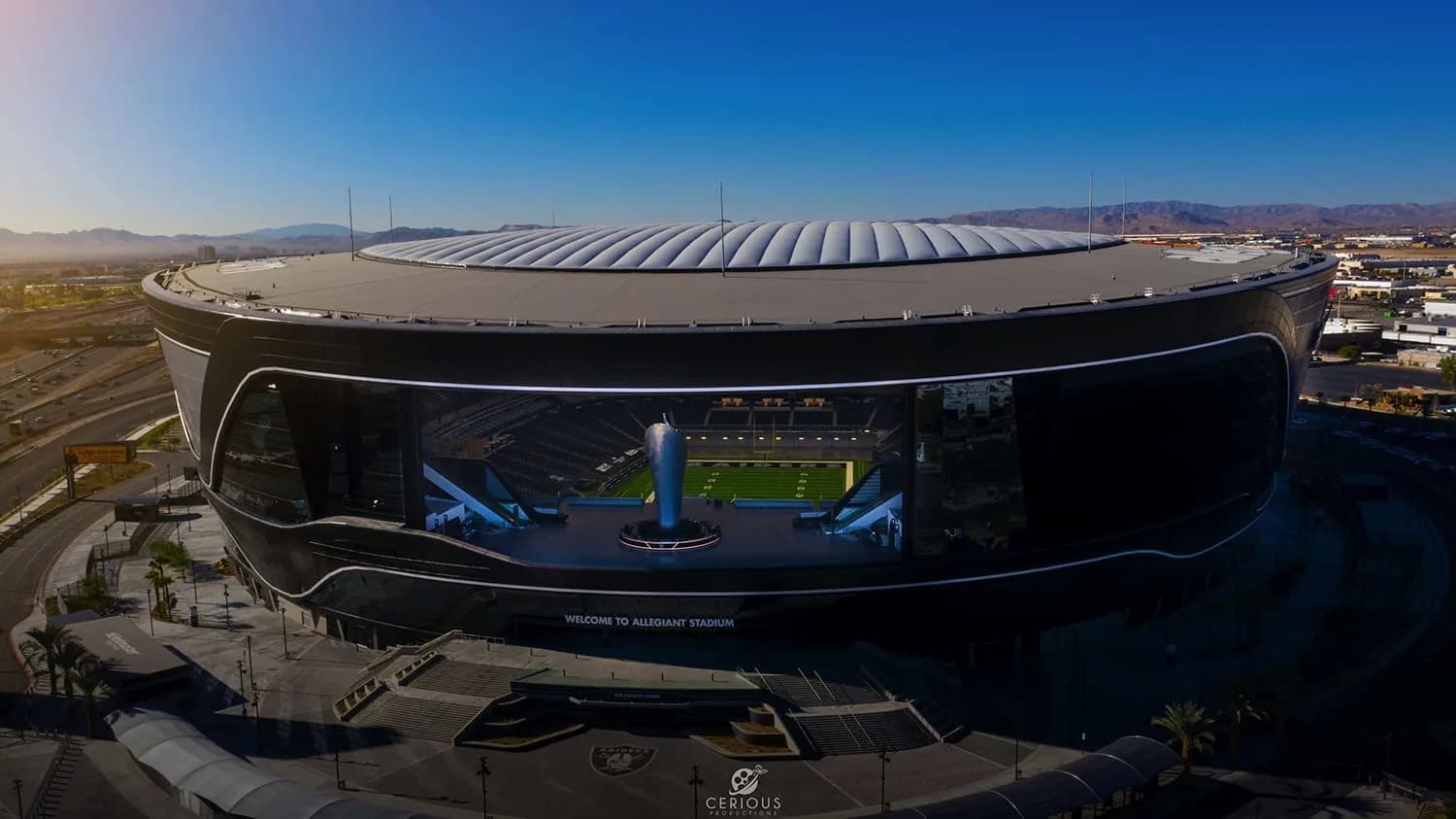 Aerial view of a curved glass-front stadium with a football field visible inside and mountains in distance