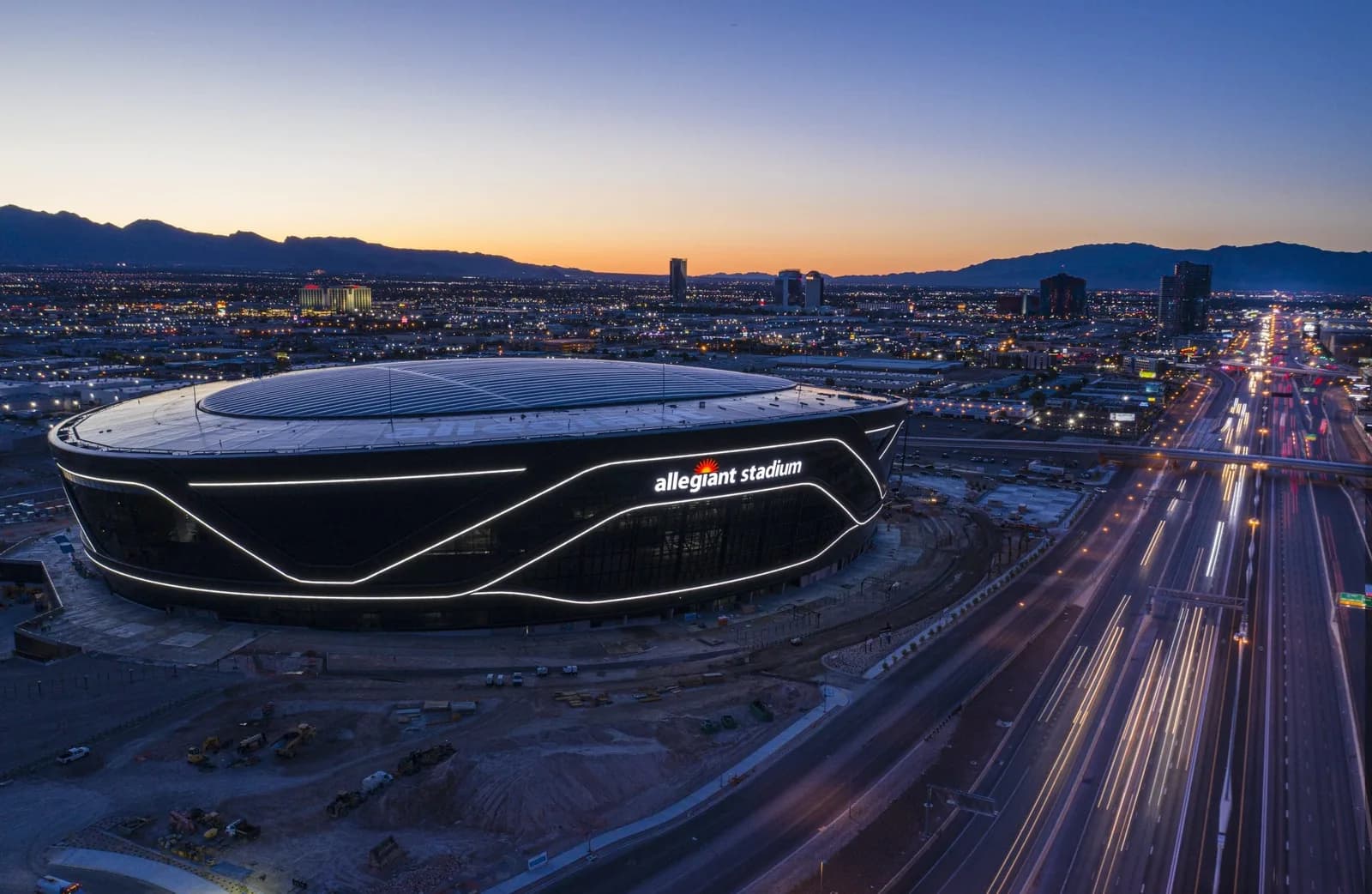 Drone view of Allegiant Stadium at dusk, curved white lines on the black facade, city lights and a highway
