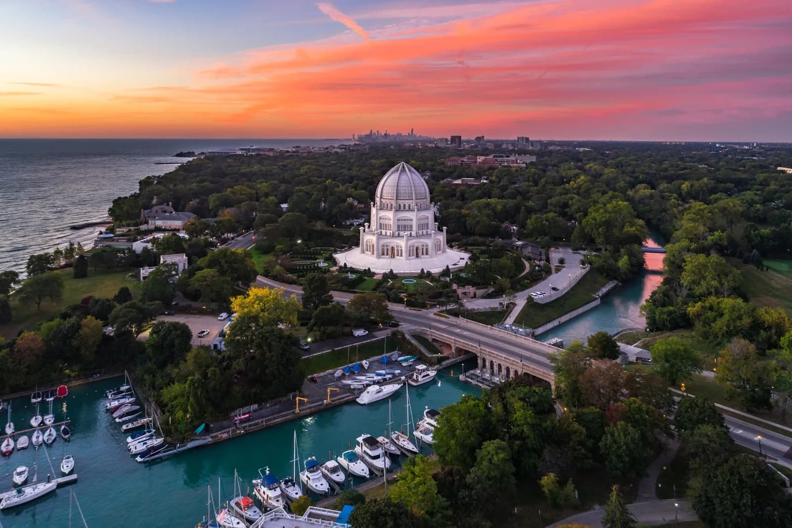 Neoclassical white domed building at center, marina with boats in foreground, sunset over trees and skyline