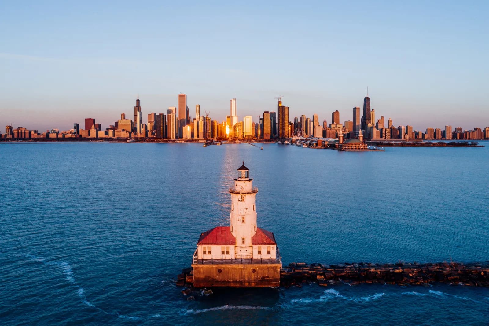 White lighthouse with red roof on a rocky jetty, blue water, and a sunlit city skyline