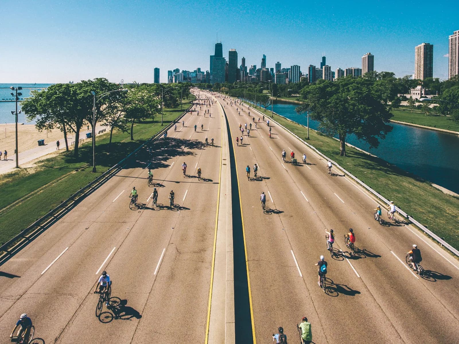 Aerial view of a divided road with cyclists on both sides, trees, a river, and a city skyline