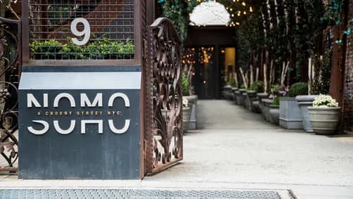 Metal gate with circular cutouts, left dark sign panel with white letters, entrance with potted plants