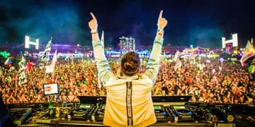 DJ on stage from behind in a white jacket with black stripe, crowd with colorful lights and large letters