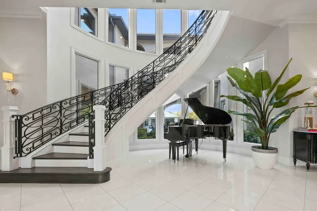 Elegant white foyer with a curved staircase, black iron railing, grand piano, and large plant