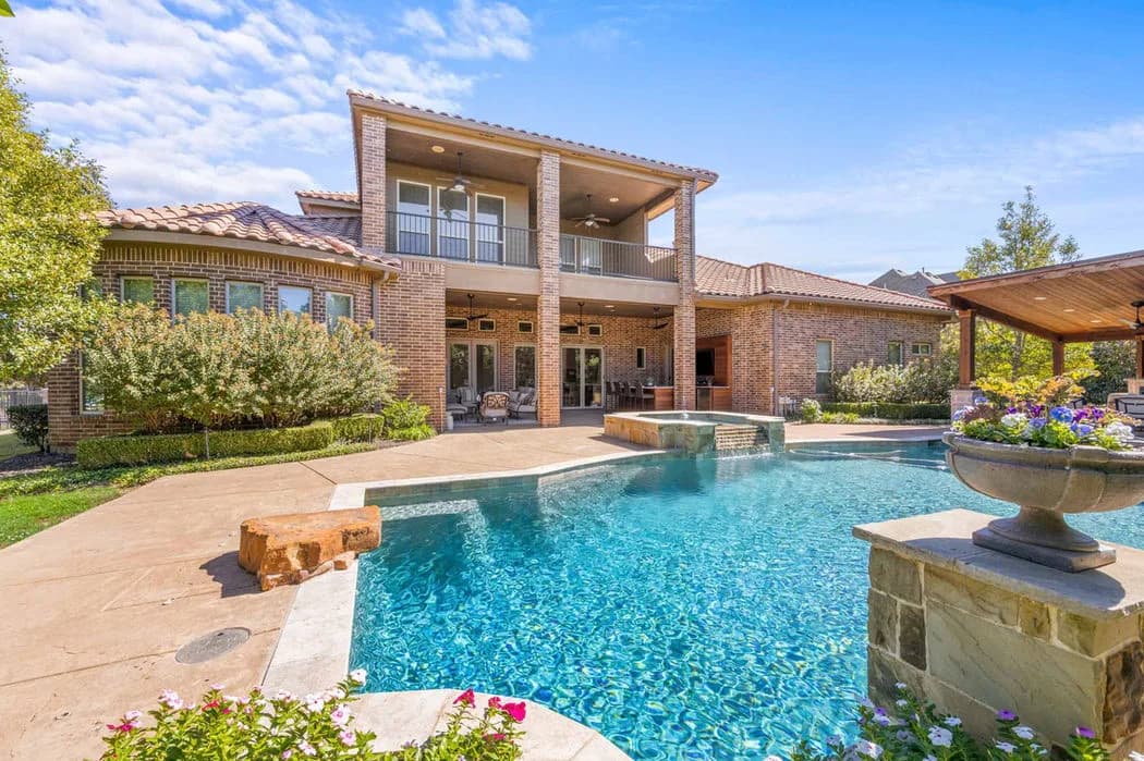 Backyard pool with a brick two-story house, balcony and covered patio; a stone urn planter with flowers