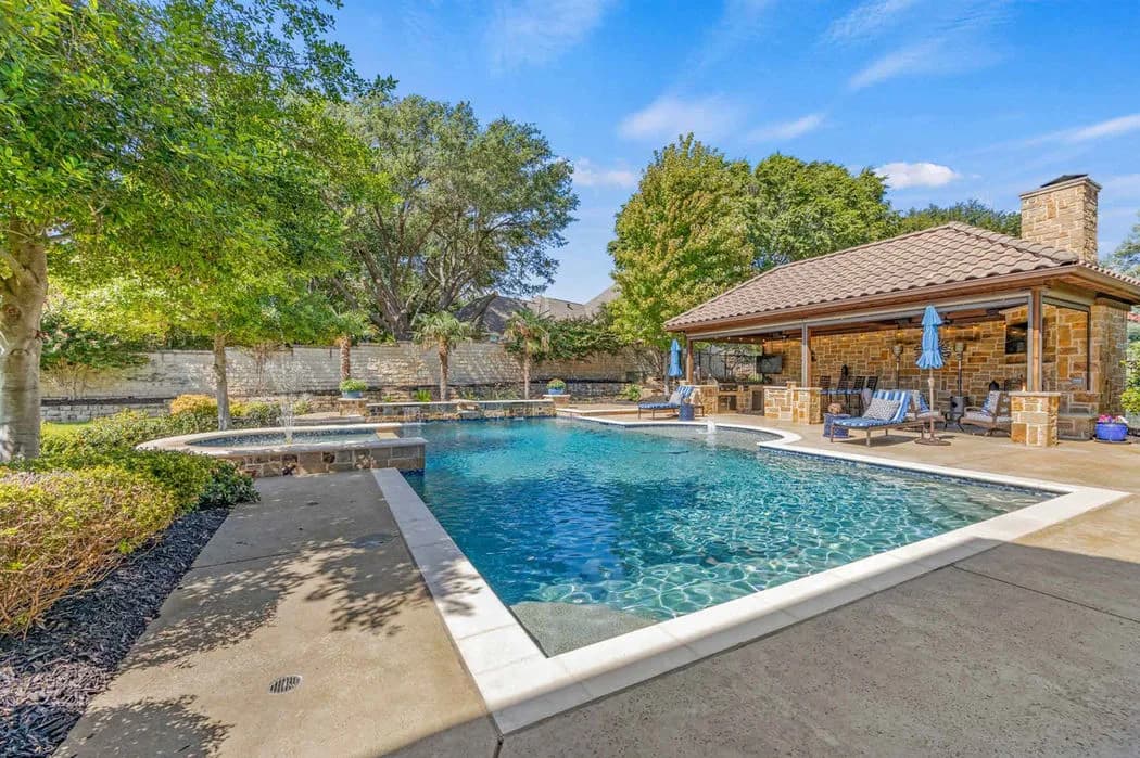 Pool area with stone pavilion and tiled roof, lounge chairs and blue umbrellas, surrounded by trees