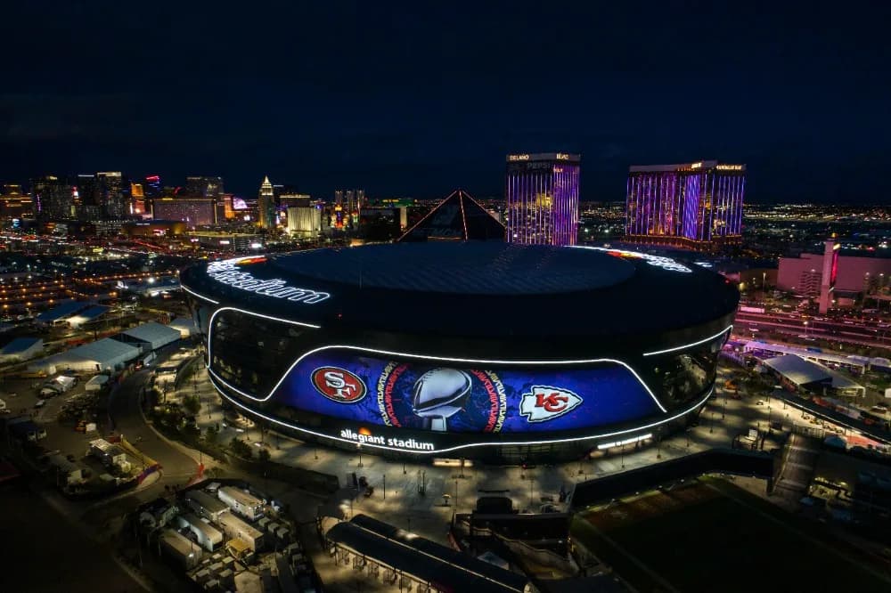 Aerial view of Allegiant Stadium at night with lit exterior displaying team logos and a helmet