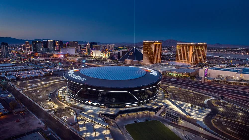 Aerial view of a circular stadium with a blue roof, surrounded by city lights and the Luxor pyramid