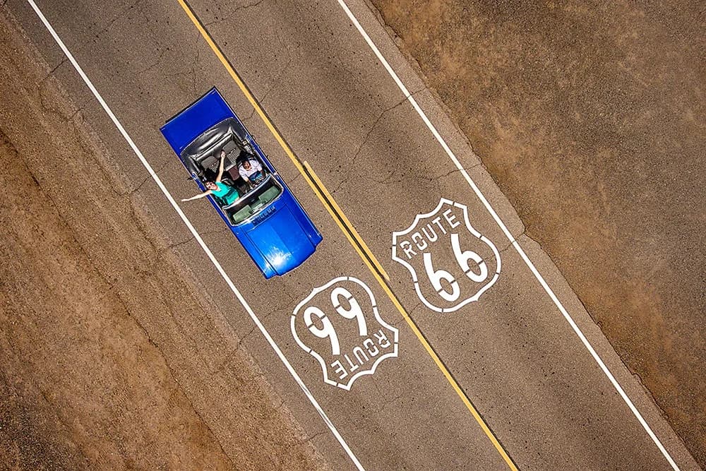 Aerial view of a blue convertible with two passengers on Route 66 shields painted on the road