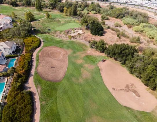 Aerial view of a golf course with two large sand bunkers, green fairways, trees, and surrounding houses