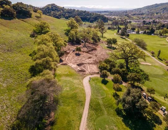 Aerial view of a winding dirt path through green hills with scattered trees and a bare dirt clearing