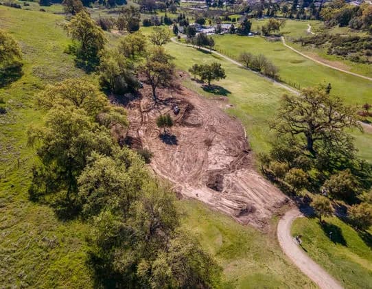 Aerial view of a green park with a cleared dirt area and surrounding trees, a curved path