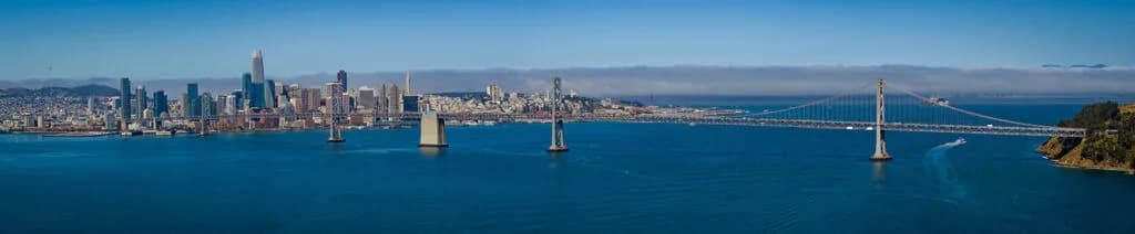 City skyline along blue water with a large suspension bridge spanning to the right