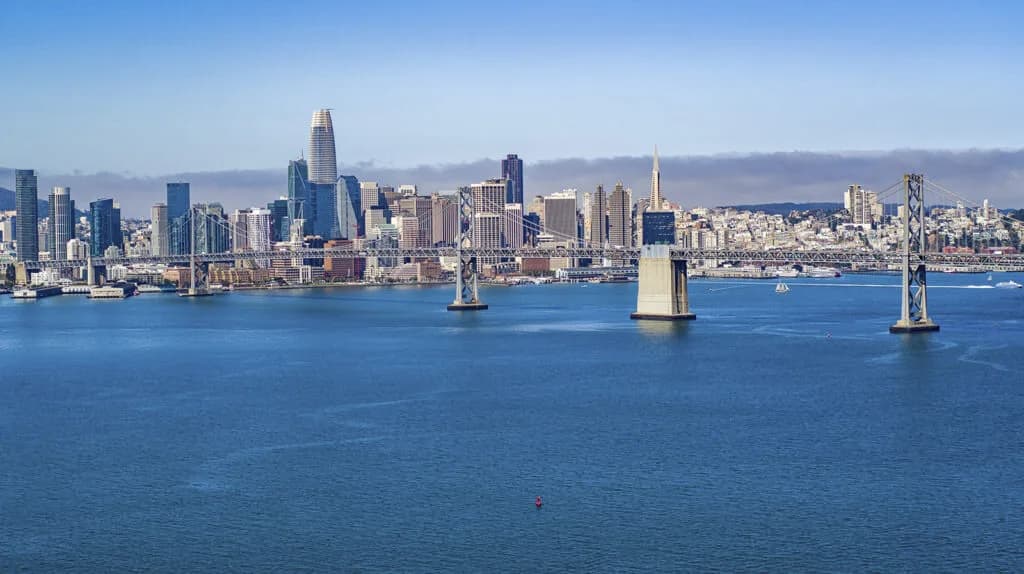 City skyline across blue water with a large suspension bridge, tall buildings, and a red buoy