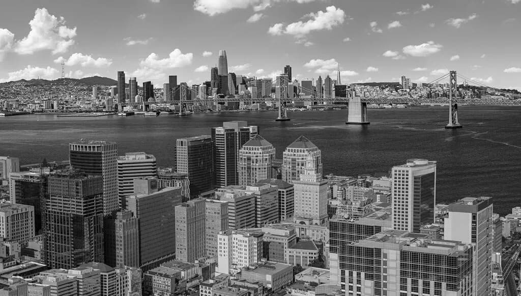 Aerial black-and-white cityscape by the water with a suspension bridge in the background