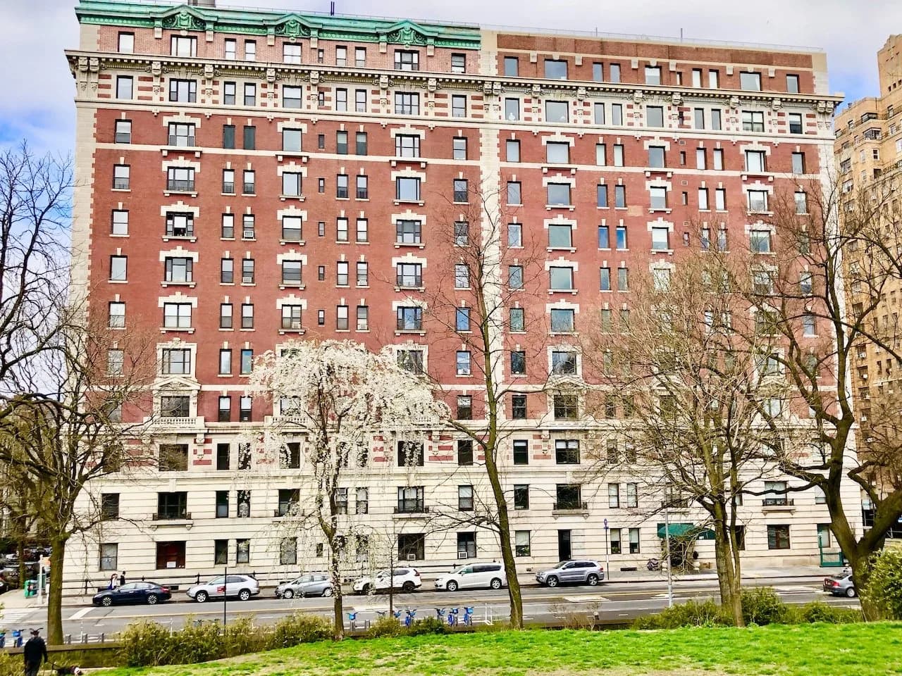 Red brick building with white trim, many windows, bare trees, and a grassy foreground with a street and cars