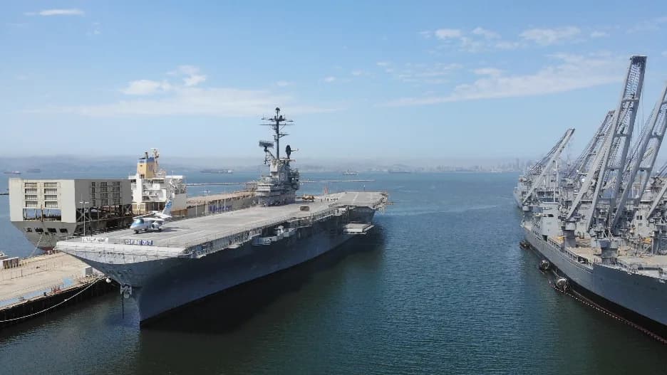 Dock scene with a large aircraft carrier at left and tall cranes on the right beside calm water