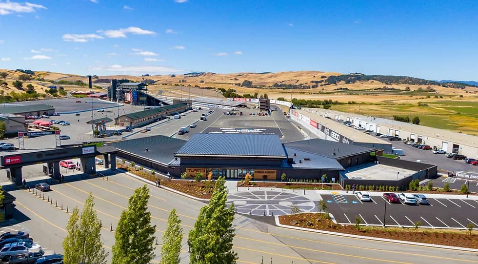 Aerial view of a race facility with a dark-roofed building, surrounding parking and hills under a blue sky