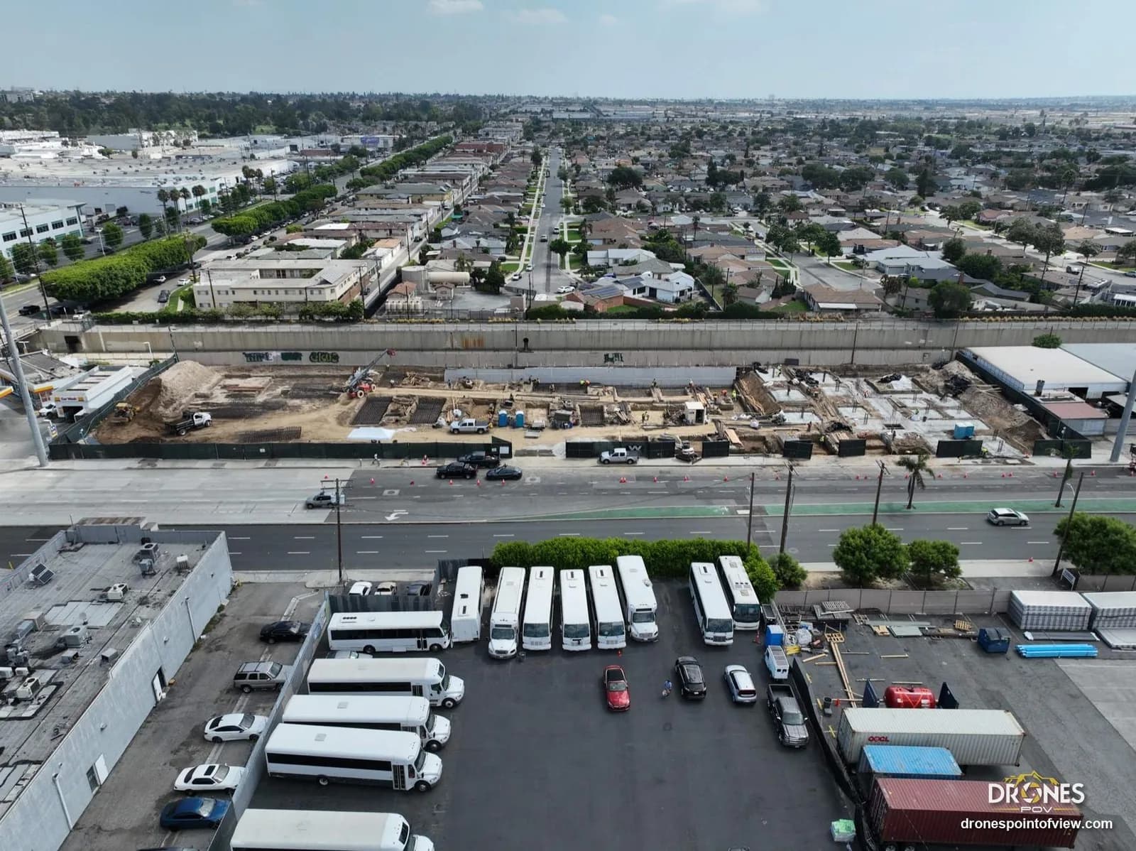 Aerial view of a construction site along a highway, with a bus yard and residential area in the distance; logo bottom right