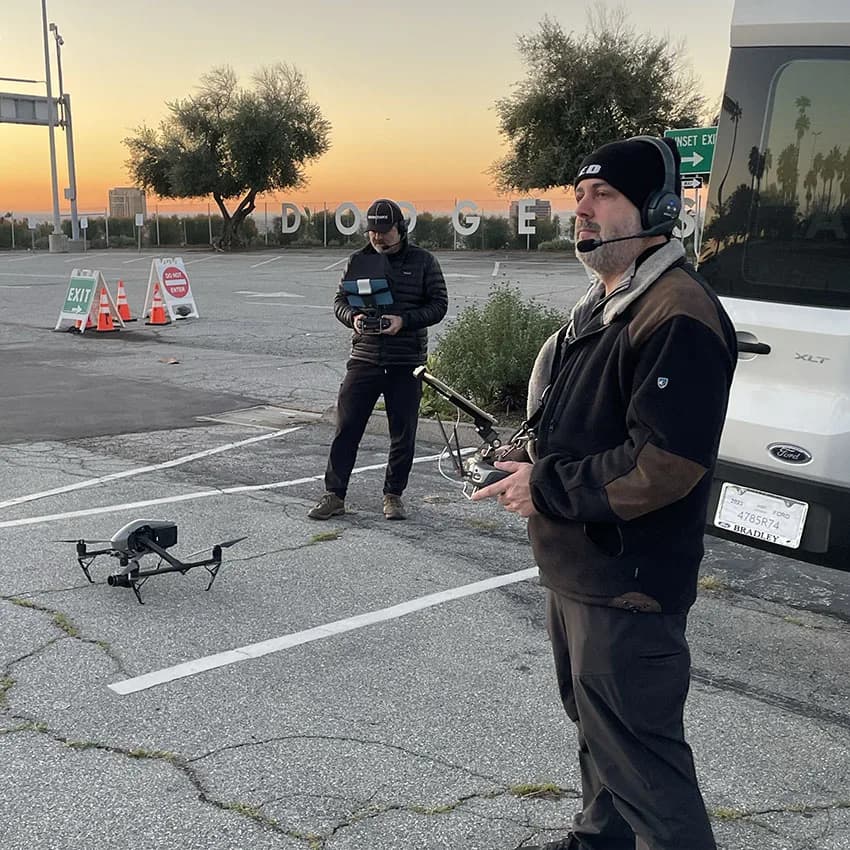 Two drone operators in a parking lot at sunset, one with headset and controller, a drone on the ground nearby