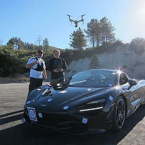 Two men with drone controllers beside a black sports car, a quadcopter flying overhead