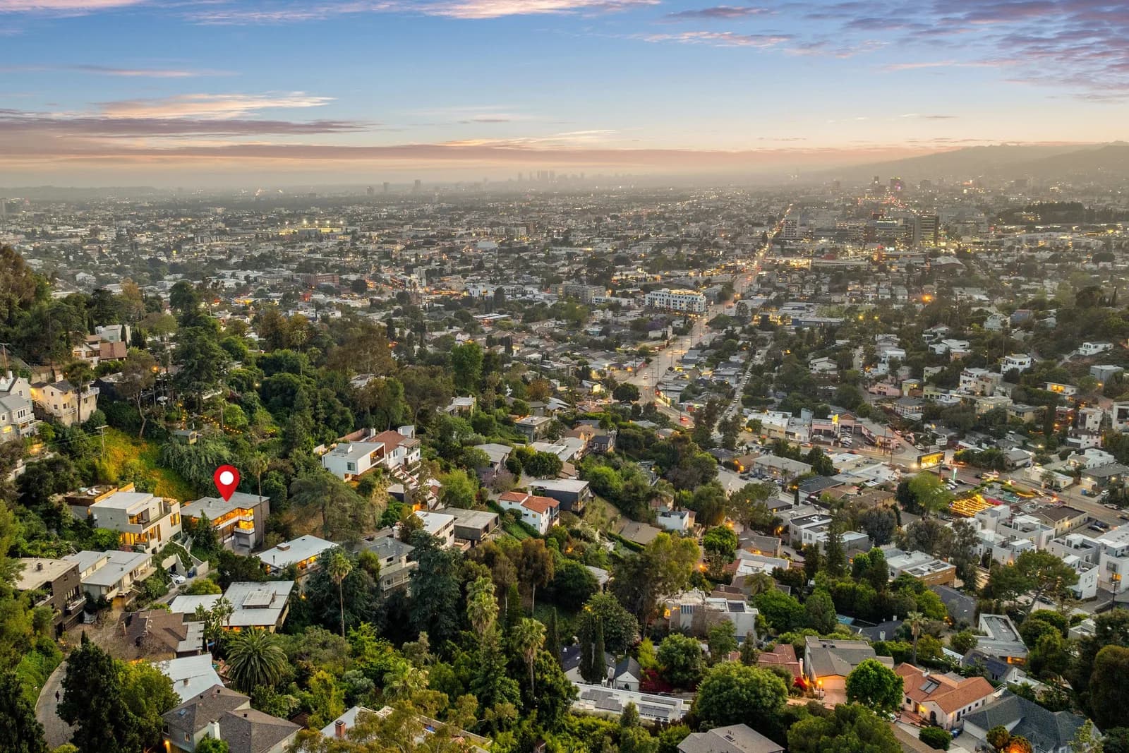 Aerial view of a hillside suburb with houses and trees, red pin marks a home, city skyline in the distance