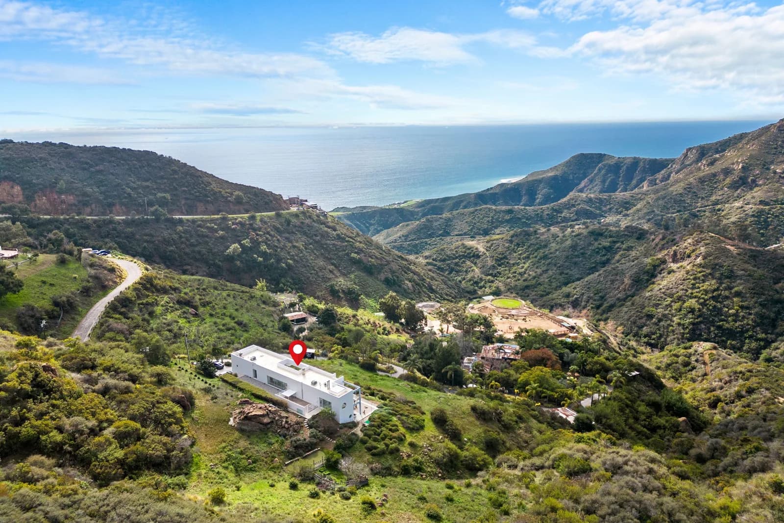 White modern house on a hillside with a red pin, green hills and ocean in the background