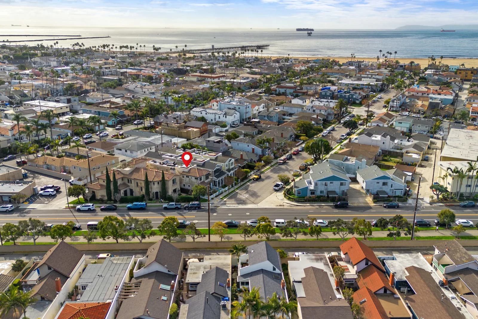 Aerial view of a coastal town with houses, palm trees, a pier, and the beach; a red pin marks a roof