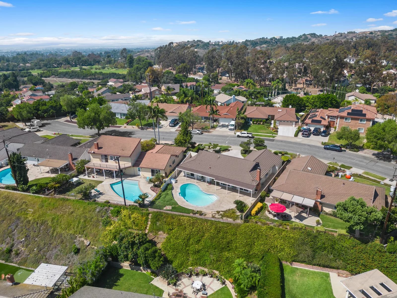 Aerial view of a suburban neighborhood with single-family homes, curved blue pools, and distant hills
