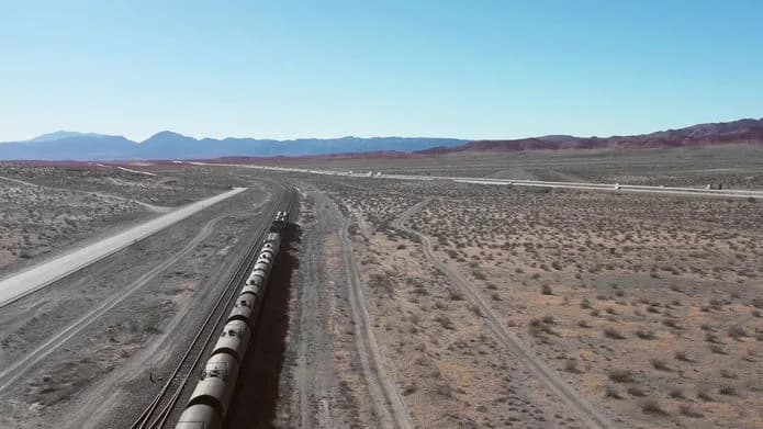 Aerial view of a long freight train along tracks in a desert with sparse shrubs and distant mountains