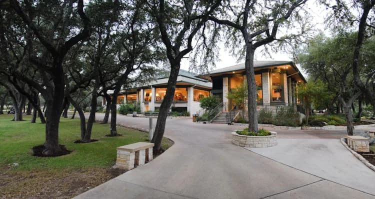 Tree-lined driveway leading to a lit, two-story building with large windows and stone planters