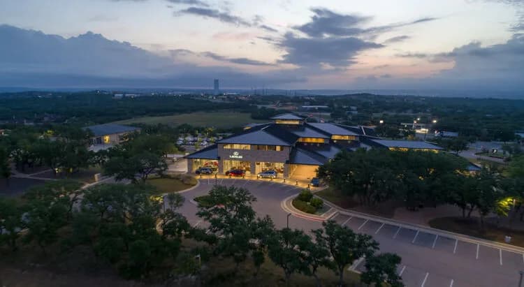 Aerial view of a modern, multi-building campus with a curved driveway, trees, and a lit parking lot at dusk