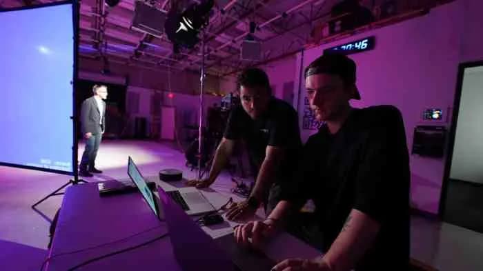 Two technicians lean over laptops at a desk in a purple-lit studio as a man speaks on a blue screen