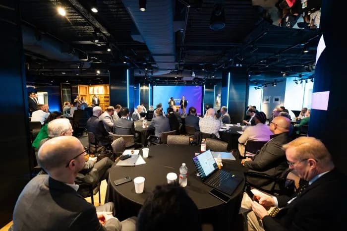 Conference room with round tables and attendees, panel on stage under a blue backdrop